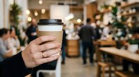 Person holding takeaway coffee cup in busy cafe with blurred customers and festive decorations in background, suitable for hospitality and beverage marketing.
