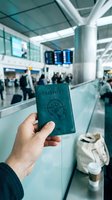Traveler holding teal passport at airport counter with flight information display and blurred passengers in background, ready for international travel and immigration check-in process.