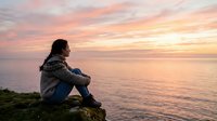 Young woman with long dark hair, wearing casual denim and a knit sweater, sitting on coastal rocks at sunset gazing toward the ocean horizon. Warm golden and pink sky reflects on calm water. Suitable for wellness, lifestyle, and contemplation themes.