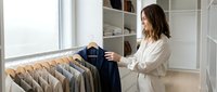 Young woman organizing clothes in modern walk-in closet with white shelving and natural light from window. Professional wardrobe management and home organization.