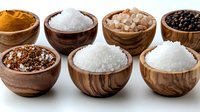 Various types of salt and spices in wooden bowls arranged in row on white background, including sea salt, black pepper, turmeric powder and pink Himalayan crystals.