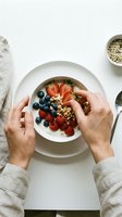 Hands holding spoon over healthy breakfast bowl with fresh berries, granola and yogurt on white table for morning nutrition and wellness lifestyle.