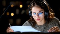 Young woman in glasses and headphones studying at night with bokeh lights in background, writing in notebook while listening to music, focused on education.
