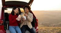 Two diverse women friends enjoying coffee break during road trip adventure sitting in car trunk at golden hour sunset sharing laughs and warm beverages outdoors.
