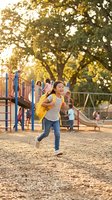 Happy young girl with yellow backpack running at school playground during golden hour with children playing in background.