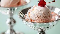 Creamy peach ice cream scoop in elegant silver dessert bowl topped with maraschino cherry, served on mint green background.