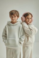 Two children in cozy cream-colored loungewear sets pose together, one wearing pink wireless headphones, creating a modern minimal lifestyle portrait against neutral background.
