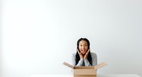 Young woman with dark hair and South Asian ethnicity, wearing business casual gray blazer, emerging from cardboard box with surprised and excited expression against white background, isolated for design use.