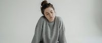 Young woman with messy bun hairstyle wearing oversized gray sweatshirt sitting peacefully with eyes closed in minimalist white studio setting for wellness content.