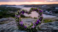 Heart-shaped floral wreath with purple wildflowers and green foliage resting on rocky cliff against dramatic sunset sky and distant water view. Romantic nature composition.