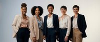 Diverse group of five professional women standing together in business casual and formal attire against neutral background, representing corporate teamwork and workplace diversity.