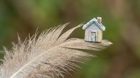 Miniature blue house with yellow door perched on brown feather against blurred natural background. Concept for home dreams and aspirations.