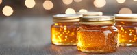 Golden honey in glass jars with metal lids on wooden surface against blurred bokeh lights background, natural organic sweetener product photography.