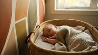Peaceful newborn baby sleeping in wicker basket with soft blanket near window with warm natural light creating cozy nursery atmosphere.