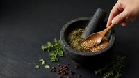 Hand grinding coriander seeds in stone mortar and pestle with fresh herbs on dark slate background for cooking and spice preparation.