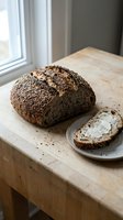 Whole grain seeded bread loaf with slice spread with cream cheese on wooden table by window with natural light for bakery and food styling.