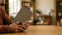 Senior adult with weathered hands holding closed book on wooden table in home library setting with bookshelves and plants.