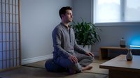 Young Caucasian man in casual gray shirt practicing mindfulness meditation sitting cross-legged on wooden floor in modern minimalist room with plants.