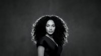 Young African American woman with voluminous curly hair in black outfit against dark background. Professional portrait photography for fashion campaigns.