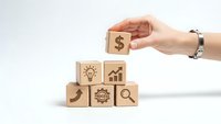 Hand placing wooden cube with dollar sign on pyramid of business icon blocks representing financial growth and investment strategy concepts.