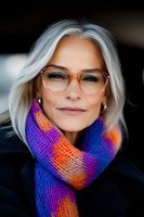 Mature woman with silver hair wearing stylish glasses and vibrant multicolored knit scarf smiles confidently at camera. Natural aging beauty portrait with soft lighting.