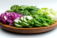 Fresh sliced vegetables on wooden plate including purple cabbage, cucumber, lettuce and green herbs arranged in rows for healthy eating and salad preparation.