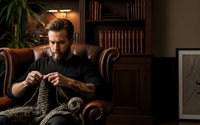 Bearded man knitting in cozy home library with leather armchair and bookshelves, relaxing hobby activity for stress relief and mindfulness practice.