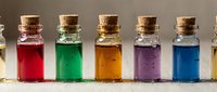 Colorful liquid-filled glass bottles with cork stoppers arranged in a row, displaying red, green, amber, purple, and blue solutions for chemistry, potion, or laboratory concepts.