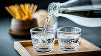 Clear liquid poured from glass bottle into two drinking glasses on wooden serving tray, with blurred background showing appetizer sticks in holder.