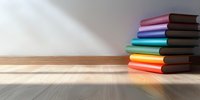 Stack of colorful hardcover books arranged in rainbow order on wooden surface against white wall with dramatic sunlight and shadow creating minimal composition.