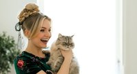 Young woman with blonde hair styled in braids, fair complexion, wearing black floral top, holding and playing with fluffy tabby cat indoors.
