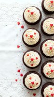 Chocolate cupcakes with vanilla buttercream frosting decorated with red heart sprinkles and colorful confetti on white wooden background for Valentine's Day.