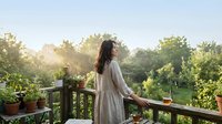 Young woman in white dress enjoying peaceful morning on wooden deck with coffee cup surrounded by lush green garden and warm sunlight streaming through trees.