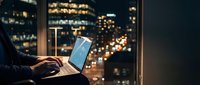 Professional working on laptop computer at night with city lights bokeh background through office window creating productive workspace atmosphere.