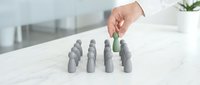 Hand selecting green cone shaped peg from row of gray pegs on white marble table, representing choice, diversity and standing out from crowd.