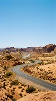 Winding asphalt road curves through arid desert landscape with sparse vegetation, rocky outcrops, and a meandering river under clear blue sky for travel and nature themes.
