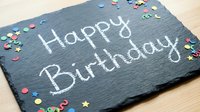 Happy Birthday written in chalk on a dark slate board surrounded by colorful confetti stars and shapes on a wooden surface.
