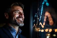 Mature bearded businessman with gray hair smiling confidently near window with blurred city lights at night, wearing navy blue blazer, side view portrait.