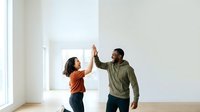 A young Black man and woman give each other a high-five in an empty, bright room with white walls and natural light from a window, celebrating success or agreement.