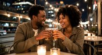 Happy African American couple toasting with drinks on romantic dinner date at outdoor restaurant with string lights creating warm evening atmosphere.