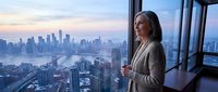 Middle-aged woman with long dark hair and light skin, wearing a beige cardigan, standing by floor-to-ceiling windows in a high-rise office, gazing at sprawling cityscape at dusk. Corporate professional environment.