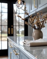 Elegant kitchen interior with dried flowers in beige ceramic vase on marble countertop, brass hardware details, and natural light through black framed windows.