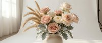 Elegant blush pink roses with dried wheat stems and dusty miller foliage in neutral ceramic vase on white surface for wedding decor and romantic arrangements.