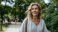 Confident middle-aged Caucasian woman with blonde curly hair smiling in park setting, wearing casual light clothing, representing wellness and positive aging.