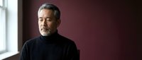 Mature Asian businessman in black turtleneck standing by window in modern office, contemplating future business decisions with confident expression.