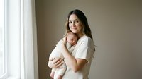 Young Caucasian mother in white shirt tenderly holding sleeping newborn baby near bright window in minimalist home interior for family lifestyle content.