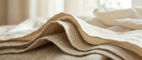 Soft beige linen fabric with natural texture draped elegantly on bed in bright bedroom interior. Organic cotton textile material for home decor and fashion design.