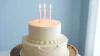 Elegant white birthday cake with pink candles and decorative bow on wooden stand against soft blue background for celebration events.
