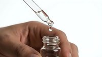Closeup of a hand holding a glass dropper bottle with a liquid drop falling, skincare serum or essential oil concept.