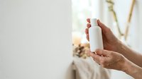 Close-up of a hand holding a white cylindrical cosmetic bottle against a blurred natural background with dried plants, suitable for skincare and beauty product marketing.
